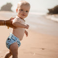Load image into Gallery viewer, A baby wearing a Reusable Swim Nappy with a whale design, enjoying a sunny day at the beach.