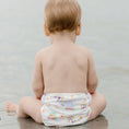Load image into Gallery viewer, Baby sitting on the beach wearing a Reusable Swim Nappy - Pastel Medley, enjoying water play.