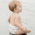 Load image into Gallery viewer, Child sitting on the beach wearing a Reusable Swim Nappy - Pastel Medley, enjoying a sunny day outdoors.