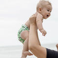 Load image into Gallery viewer, Baby wearing a Reusable Swim Nappy being held by an adult at the beach, perfect for fun and comfort.