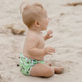 Load image into Gallery viewer, Baby sitting on the beach wearing a green Reusable Swim Nappy featuring Cockatoos, perfect for beach days.