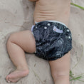 Load image into Gallery viewer, Infant wearing a Black Flora reusable swim nappy on the beach, showcasing floral design and comfort for water play.