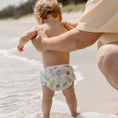 Load image into Gallery viewer, Baby wearing a Reusable Swim Nappy with a tropical pattern, standing by the water on a beach.