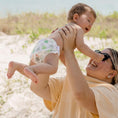 Load image into Gallery viewer, Happy baby wearing a Reusable Swim Nappy held by an adult on a beach, enjoying a sunny day outside.